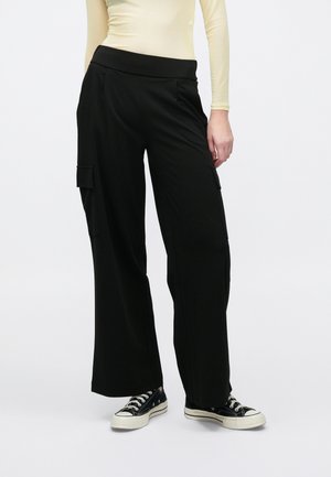Person trägt schwarze, weit geschnittene Cargohosen mit hoher Taille und schwarze flache Sneakers vor einem schlichten, hellen Hintergrund.
