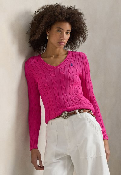 Ροζ πουλόβερ με πλεκτή υφή και V-λαιμόκοψη, με μακριά μανίκια και λογότυπο σε επιδιορθωμένο σχέδιο, σε συνδυασμό με λευκό παντελόνι με ψηλή μέση και διακοσμητική ζώνη.