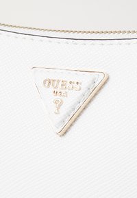 Un matériau texturé blanc avec un logo triangulaire Guess USA sur une étiquette en relief blanche, entouré d'accents en métal doré.