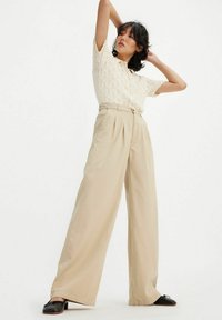 Beige weit geschnittene Hosen mit Falten, kombiniert mit einem cremefarbenen Lace-Stricktop. Die Schuhe sind schwarze Slipper. Einfaches, klares Design.