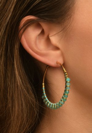 Pendientes de aro con cuentas de turquesa y acentos de alambre dorado, colgando de una oreja perforada, mostrando una textura de piel suave.
