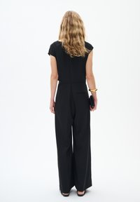 Combinaison noire à manches courtes, jambes larges et taille ajustée. Dotée d'une poche arrière et d'une texture lisse. Portée avec une pochette noire et un bracelet doré.