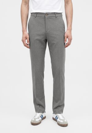 Homme portant un pantalon gris sur mesure et des baskets blanches avec des bandes bleues, debout devant un fond uni.