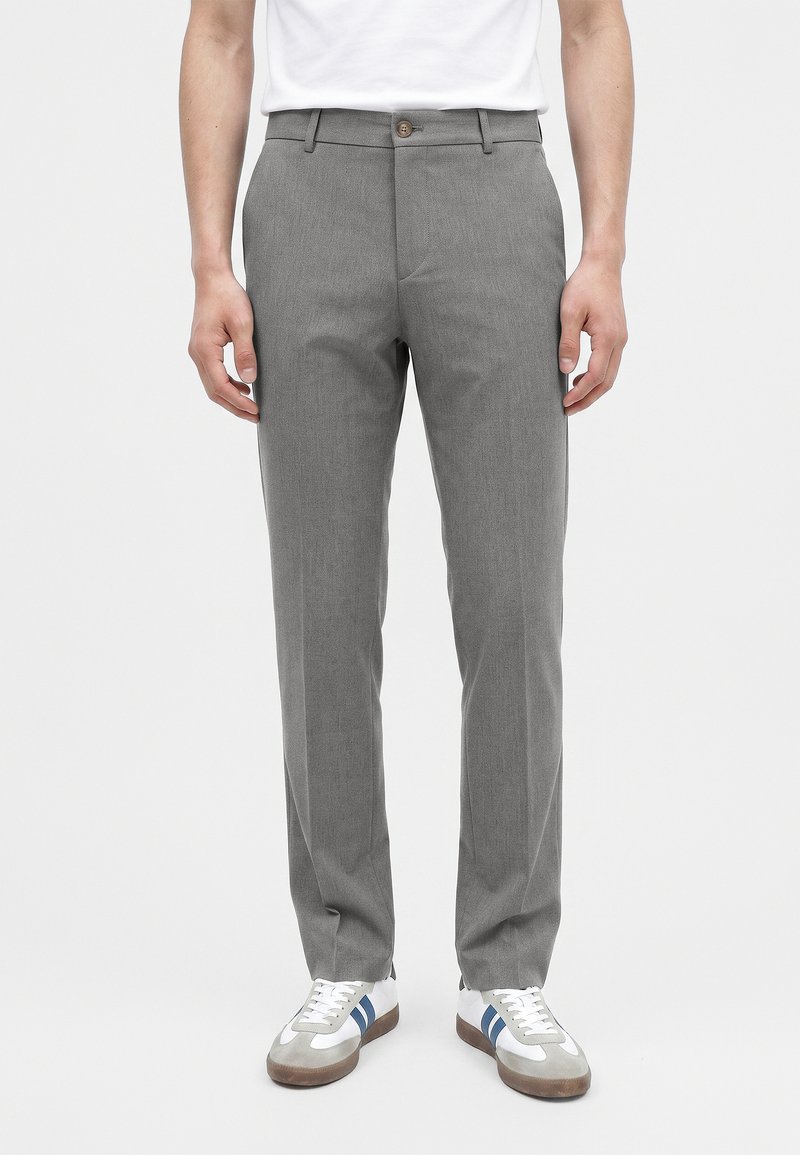 Homme portant un pantalon gris sur mesure et des baskets blanches avec des bandes bleues, debout devant un fond uni.