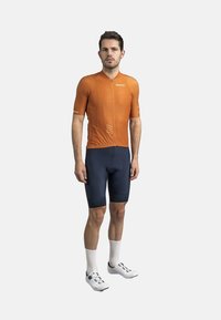 Fietsjersey in oranje met een rits aan de voorkant, gecombineerd met marineblauwe shorts. De outfit heeft ademend materiaal en een strakke pasvorm. Witte fietsschoenen.
