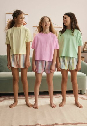 Trzy bose dziewczynki stoją w pomieszczeniu, ubrane w pastelowe koszulki i krótkie spodenki w wzory, uśmiechają się i patrzą na siebie.