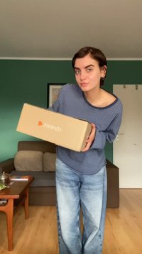 Cutie de carton maro cu text alb și un logo, ținută în mâini, purtând o cămașă cu dungi albastre și blugi decolorati.