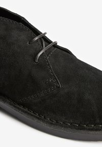 Schwarzer Veloursleder-Schnürschuh mit sichtbaren Nähten und runder Spitze, Nahaufnahme auf weißem Hintergrund.