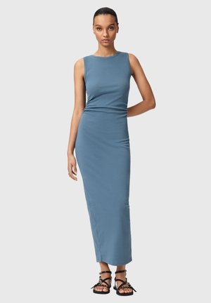 Kobieta stoi, ubrana w dopasowaną, bezrękawnikową, niebieską sukienkę do kostek oraz czarne, płaskie sandały na paski, na jasnym, jednolitym tle.