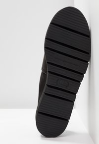 Semelle de botte noire avec de larges rainures horizontales, positionnée à la verticale contre un mur d’angle clair et un sol.