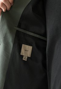 Main tenant un blazer noir ouvert montrant une doublure noire texturée avec une étiquette beige indiquant "NEXT" et la taille "M" à l'intérieur.