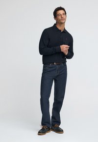 Marinblå pikétröja med långärmar och en röd logga, kombinerad med mörkblå denimjeans. Svarta skor med orange snören och ett brunt bälte.