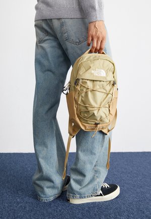Osoba w jasnoniebieskich dżinsach i czarnych trampkach trzyma mały beżowy plecak The North Face za górną rączkę.