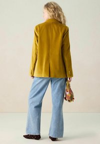Blazer en velours jaune moutarde avec un col cranté, une fente au dos, et des poignets à trois boutons, associé à un jean large bleu clair.