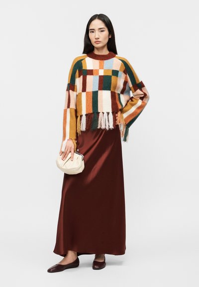 Femme aux longs cheveux bruns portant un pull multicolore à rayures avec franges, une jupe longue en satin bordeaux, des ballerines marron, et tenant un petit sac blanc.