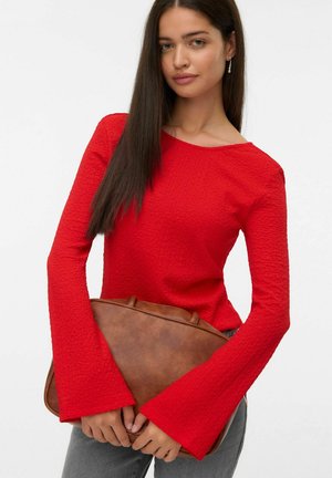 Vrouw met lang, donker haar, gekleed in een rode, gestructureerde top met lange mouwen, houdt een grote, bruine leren handtas vast tegen een effen achtergrond.