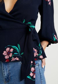 Marinebluse med indpakningsdesign, der har blomsterprint i pink og grøn. Lange ærmer med klokkeformede manchetter og en knyttet talje detalje.