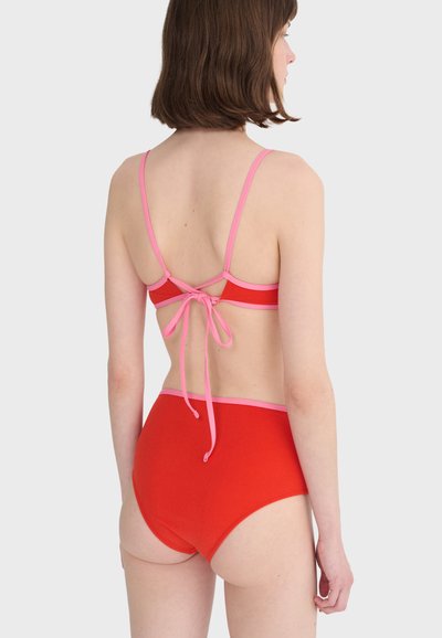 Femme aux cheveux courts bruns portant un bikini rouge avec des bordures roses, vue de dos, noué par un nœud au centre du dos.