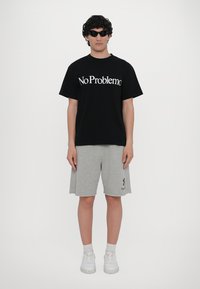 Svart t-skjorte med teksten "No Problemo" i hvitt, grå shorts, hvite joggesko og solbriller. Uformelt design uten mønstre eller dekorasjoner.