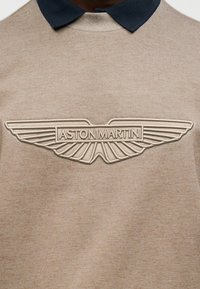 Μπεζ πουλόβερ με μπλε σκούρο γιακά, διακοσμημένο με ανάγλυφο μπεζ λογότυπο Aston Martin στο κέντρο, που παρουσιάζει καθαρές γραμμές και ομαλή υφή.