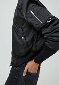 Blouson aviateur noir avec une finition brillante, des poignets côtelés, des poches latérales et une poche zippée sur la manche. Tissu lisse avec une texture subtile.