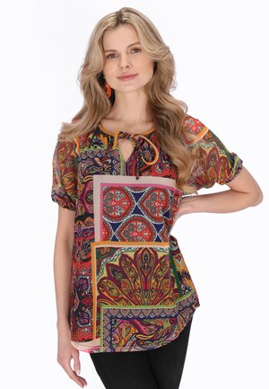 Szőke nő színes paisley és virágmintás blúzban, rövid ujjakkal, kulcslyuk nyakkivágással és megkötővel, fekete nadrágban, kezét a csípőjén tartva.