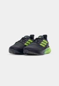 Chaussures de sport noires avec des accents verts, tige en mesh, semelle texturée et logo à trois bandes proéminent. Amortissement léger au niveau de la semelle.