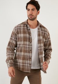 Camicia a quadri marrone e bianca in un tessuto morbido, con un botton-down sul davanti e maniche lunghe. Indossata sopra una maglietta bianca.