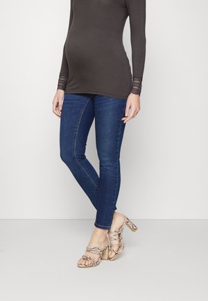 Kvinde iført mørkebrun langærmet top med blonde manchetter, tætsiddende blå jeans og hælede sandaler med slangeprint, stående mod en ensfarvet hvid baggrund.