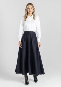 Kobieta w białej koszuli zapinanej na guziki, granatowej spódnicy midi z wysokim stanem i czarnych, spiczastych botkach za kostkę, stojąca na jednolitym tle.