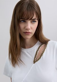 White cotton t-shirt featuring a unique neckline with a gold-tone embellished detail. Short sleeves and a fitted design.