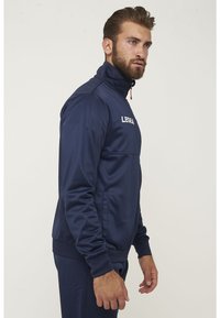 Chaqueta de chándal navy hecha de tela suave, con un alto cuello, cierre con cremallera en la parte delantera, bolsillos laterales y un logo en el pecho.