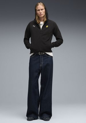 Zwarte hoodie met ritssluiting en capuchon, voorzien van een geel Ferrari-logo. Draagt losse donkere spijkerbroek en zwarte schoenen.