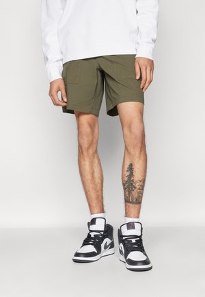 Oliivroheline cargo-lühikesed püksid koos kahe esipocketiga, kantud mustade ja valgete kõrge säärega tossudega, mille vasakul jalal on puu tätoveering.