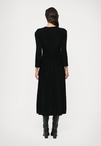 Vestido preto de manga longa com textura canelada, corpete justo e saia rodada, combinado com botas pretas até ao joelho e uma superfície lisa.