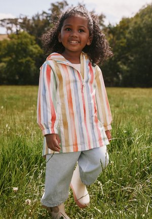 Naeratav laps lokkis juustega seisab rohelises viljapeenras, seljas mitmevärvilise triibulise jakk, heledad püksid ja kreemjad saapad.