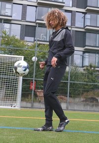 Atleet in zwart Puma trainingspak met grijze accenten, staand op groen kunstgras, jonglerend met een voetbal, met zwarte voetbalschoenen met witte details.