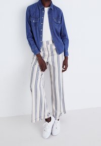 Denim shirt with two chest pockets, white ribbed top, striped wide-leg trousers in cream and navy, and white sneakers.