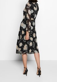 Svart blommig klänning med genomskinliga långärmar, med beige och grå blomster mönster, knä-lång fåll och figurnära midja, bärs med svarta klackar.