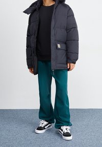 Persona vestida con pantalones turquesa, chaqueta acolchada negra con un gran bolsillo, sudadera negra y zapatillas en blanco y negro, de pie sobre una alfombra azul.