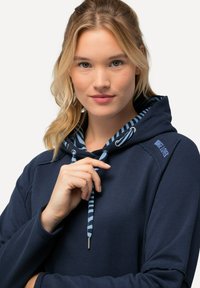 Hoodie marine avec une doublure rayée bleu clair, fermeture à cordon, et poignets côtelés. Présente un logo brodé sur l'épaule. Texture douce.