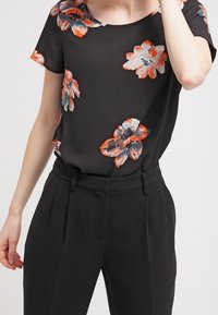 Černá halenka s krátkým rukávem s živými oranžovými a modrými květinovými vzory; doplněna o černé kalhoty s vysokým pasem a střihnutím. Hladká textura látky.