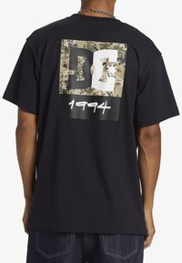 Black cotton t-shirt featuring a camo graphic with "DC" logo and "1994" in white. Short sleeves, relaxed fit, and soft texture.