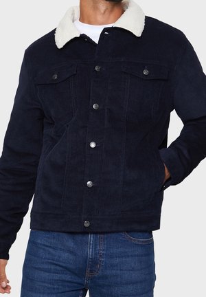 Homme portant une veste en velours côtelé bleu marine avec col sherpa blanc, fermeture par boutons, poches poitrine et jean bleu, main dans la poche sur fond uni.