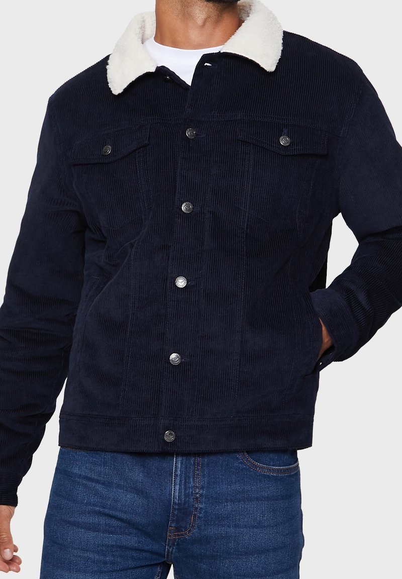 Homme portant une veste en velours côtelé bleu marine avec col sherpa blanc, fermeture par boutons, poches poitrine et jean bleu, main dans la poche sur fond uni.