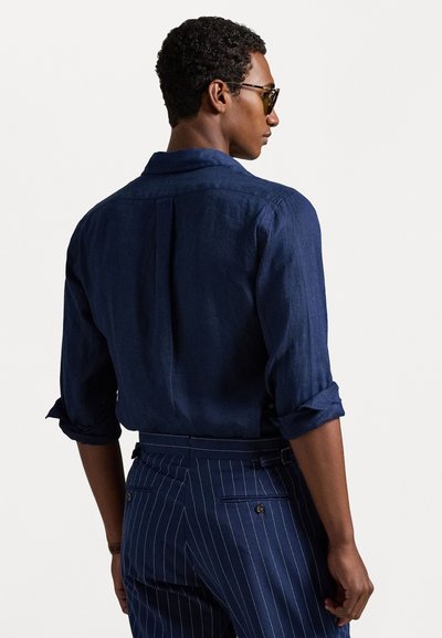 Tumši zils lina krekls ar sarullētām piedurknēm, ar klasisku apkakli un plecu detaļu aizmugurē, kombinēts ar svītrainām biksēm.