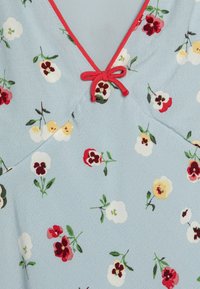 Tissu bleu clair avec un motif texturé, présentant des designs floraux rouges, blancs et jaunes. Accentué par un nœud rouge au niveau du col.