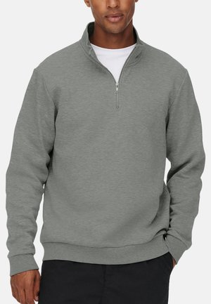 Mann trägt ein hellgraues Pullover-Hemd mit Viertel-Reißverschluss über einem weißen Hemd, mit einer Hand in der Tasche schwarzer Hosen.