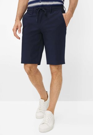 Man draagt marineblauwe korte broek met trekkoord in de taille, een patroonhemd in de broek gestopt, en witte sneakers, staat met één hand in de zak.