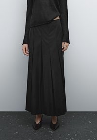 Zwarte plissé maxi rok van lichtgewicht stof, gecombineerd met een zwart langemouwen top en zwarte puntige hakken op een textuurvloer.